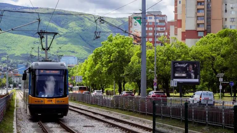 Tramwaj miejski jadący przez miasto, widoczny na tle budynków oraz zielonych drzew.