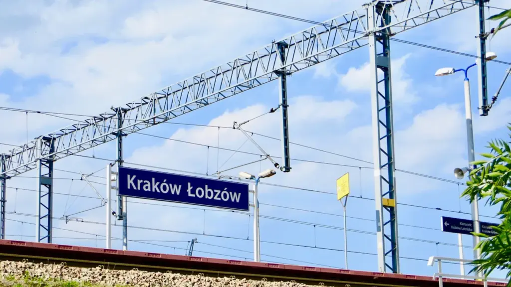 Tablica informacyjna stacji kolejowej Kraków Łobzów na tle słonecznego nieba i sieci trakcyjnej